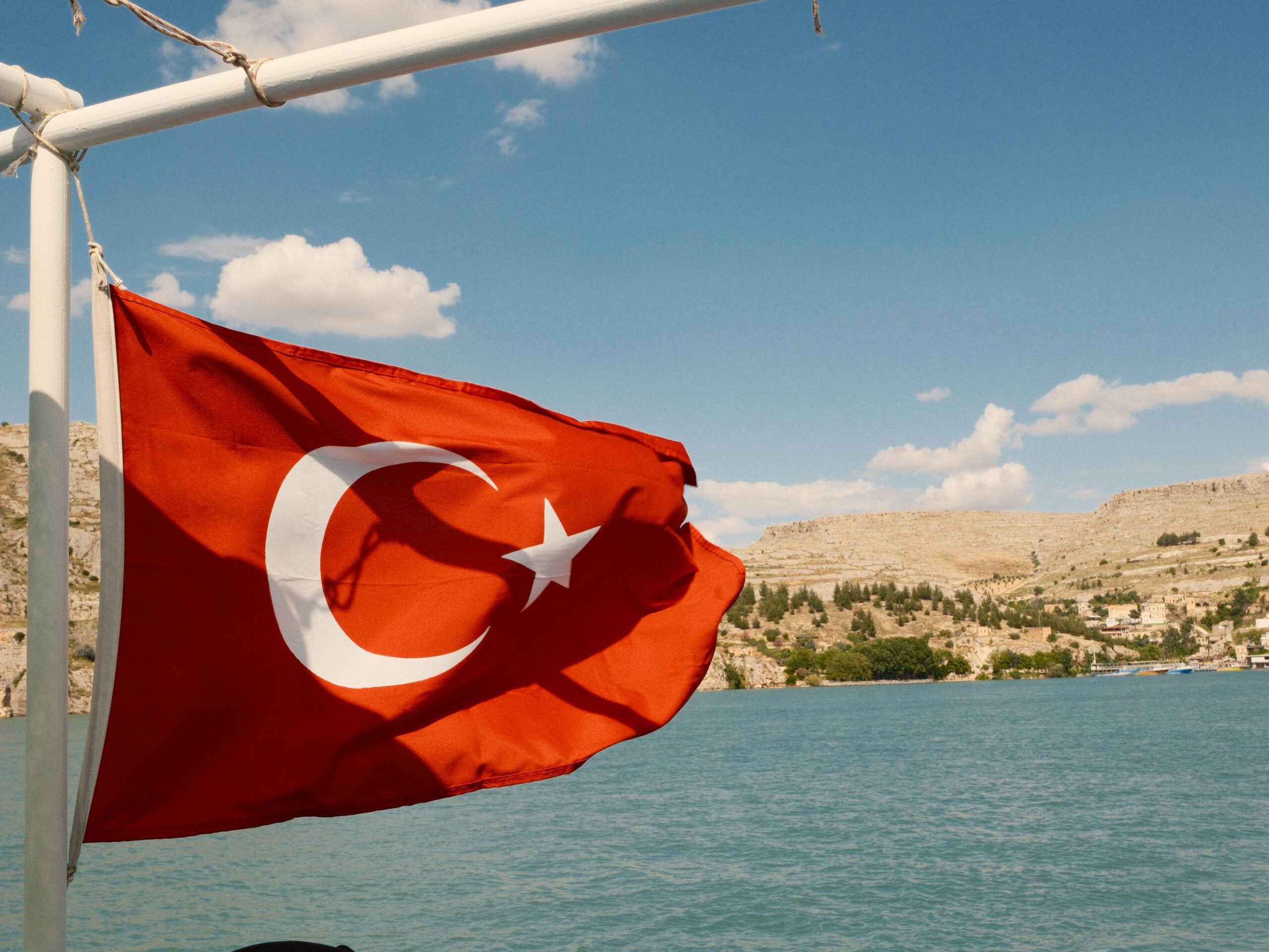 Turkish flag flying from a boat with a scenic landscape of water and mountains in the background, representing Turkish national identity